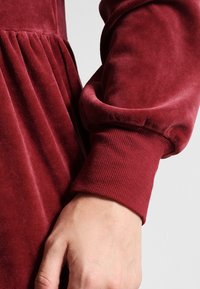 Bordowa sukienka z długim rękawem o miękkiej, welurowej teksturze i ściśniętych mankietach. Materiał elegancko opada z zgromadzonej talii.