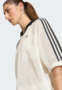 Biela sieťovina mikina s čiernym golierom, krátkymi rukávmi s tromi čiernymi pruhmi a malým logom na hrudníku.