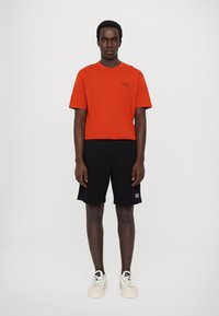 Tricou roșu din bumbac cu mâneci scurte și un logo mic, purtat cu șorturi negre. Modelul poartă adidași albi. Fundal simplu.