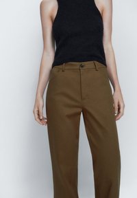 Black sleeveless top and brown tailored trousers. Top is ribbed, fitted; trousers are high-waisted with a button closure and side pockets.