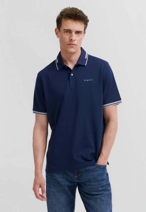 Giovane uomo che indossa una polo blu navy con bordi bianchi su colletto e maniche, abbinata a jeans blu, in piedi davanti a uno sfondo semplice.