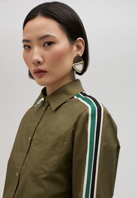 Olive green shirt with a button-up design, featuring contrasting black, white, and teal stripes on the sleeves. Large statement earrings displayed.