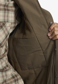 Brun jacka med en mjuk fleecefoder och en innerficka. Utsidan har en slät yta, och det finns rutiga detaljer synliga.