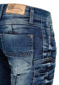 Jean en denim avec une finition bleu délavé, présentant une patch en cuir étiqueté "Cipo & Baxx", un design à cinq poches et des détails de couture décorative.