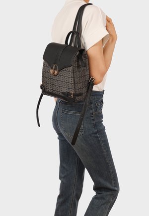 Donna che indossa jeans blu e una maglietta bianca a maniche corte, con uno zaino nero con motivo, patta in pelle e fibbia dorata, portato su una spalla.