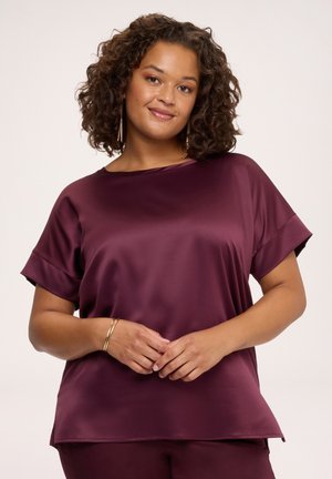 Donna con capelli ricci che indossa una maglietta bordeaux in raso a maniche corte e pantaloni abbinati, sorride dolcemente con le mani giunte davanti.