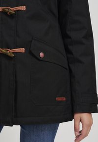 Svart ytterjakke laget av teksturert materiale, med brune tau-lukkinger, en sidelomme og en metalllogo-detalle.