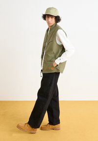 Olive green vest with multiple pockets over a white shirt, paired with black pants featuring floral patterns and brown boat shoes.