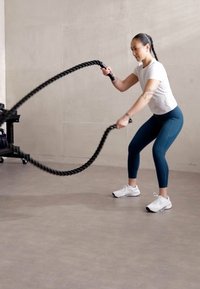 Nő fekete harci kötelekkel edz egy konditeremben. Fehér pólót, türkiz színű leggingset és fehér sportcipőt visel. Minimalista háttér.