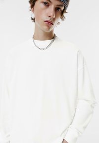Camicia bianca a maniche lunghe con scollo rotondo e vestibilità rilassata. Presenta una collana in catena argentata e un berretto a righe. Tessuto liscio.