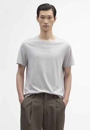 Jeune homme aux cheveux raides et foncés portant un t-shirt gris clair uni et un pantalon plissé marron foncé, debout les mains dans les poches.