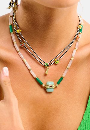 Collier superposé avec des perles vertes, roses et noires, orné d'une pierre centrale bleu clair et d'accents dorés, mettant en valeur des textures variées.