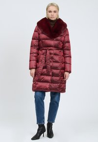 Vrouw in een lange bordeauxrode pufferjas met een bontkraag, een vaste riem, blauwe jeans en zwarte enkellaarsjes met hak tegen een neutrale achtergrond.