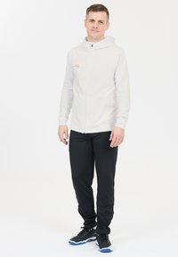Lettet, hvid lynlås hoodie med hætte, parret med sorte bukser. Bemærkelsesværdige detaljer inkluderer et lille guldlogo og atletiske sorte sko.