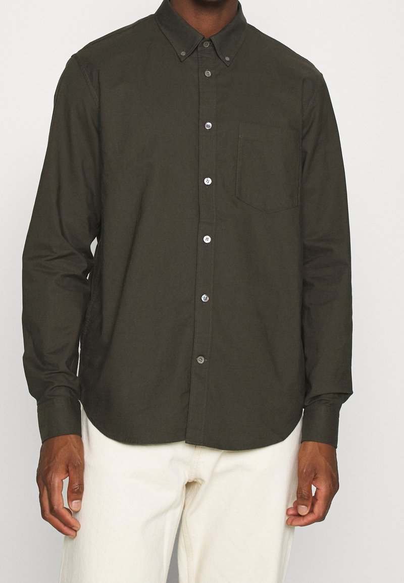 Camicia abbottonata verde scuro con colletto, maniche lunghe e taschino sul petto; presenta un orlo arrotondato e bottoni chiari a contrasto.