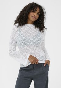 Hvid blondetop med lange ærmer og rund halsudskæring, prydet med et blomstermønster og klokkeærmer, parret med grå skrædderbukser.