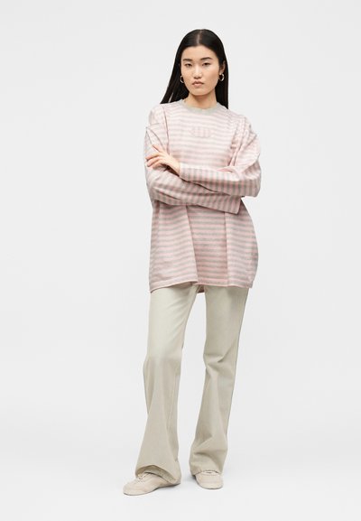 Νεαρή γυναίκα στέκεται με σταυρωμένα χέρια φορώντας μπλούζα μακριών μανικιών με ροζ και γκρι ρίγες και μπεζ φαρδιά παντελόνια με αθλητικά παπούτσια.
