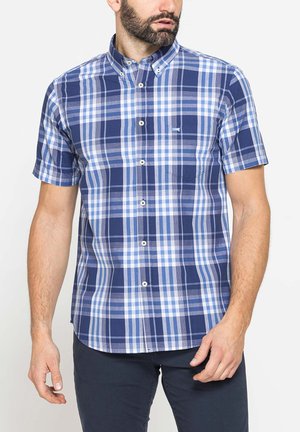 Uomo che indossa una camicia a quadri blu e bianca a maniche corte con taschino frontale, abbinata a pantaloni scuri.
