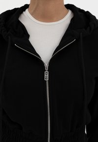 Mustat vetoketjuhupparit, joissa on pehmeä rakenne, hopeiset vetoketjut, kerroksellinen huppu ja joustava vyötärö. Kerroksena valkoisen urheilupaidan päällä.