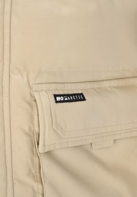 Bézs dzseki, melyen feltűnő zsebfedő található, sima textúrával és fekete "MOARCTIC" címkével, amely a zsebre van varrva. Testhezálló szabás.