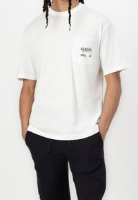 Biały bawełniany t-shirt z krótkimi rękawami, z kieszenią na piersi i czarnym nadrukiem z napisem "ODESSA" z detalem w kratkę.