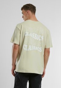 Gai beigā kokvilnas T-krekls ar lieliem baltiem burtiem "URBAN CLASSICS" aizmugurē. Īsas piedurknes, brīvs piegriezums, patīkama tekstūra.
