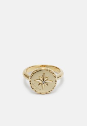 Gouden ring met een ronde voorkant met een verhoogde achtpuntige ster en een kleine heldere edelsteen in het midden, gegraveerd met het woord "GUESS".