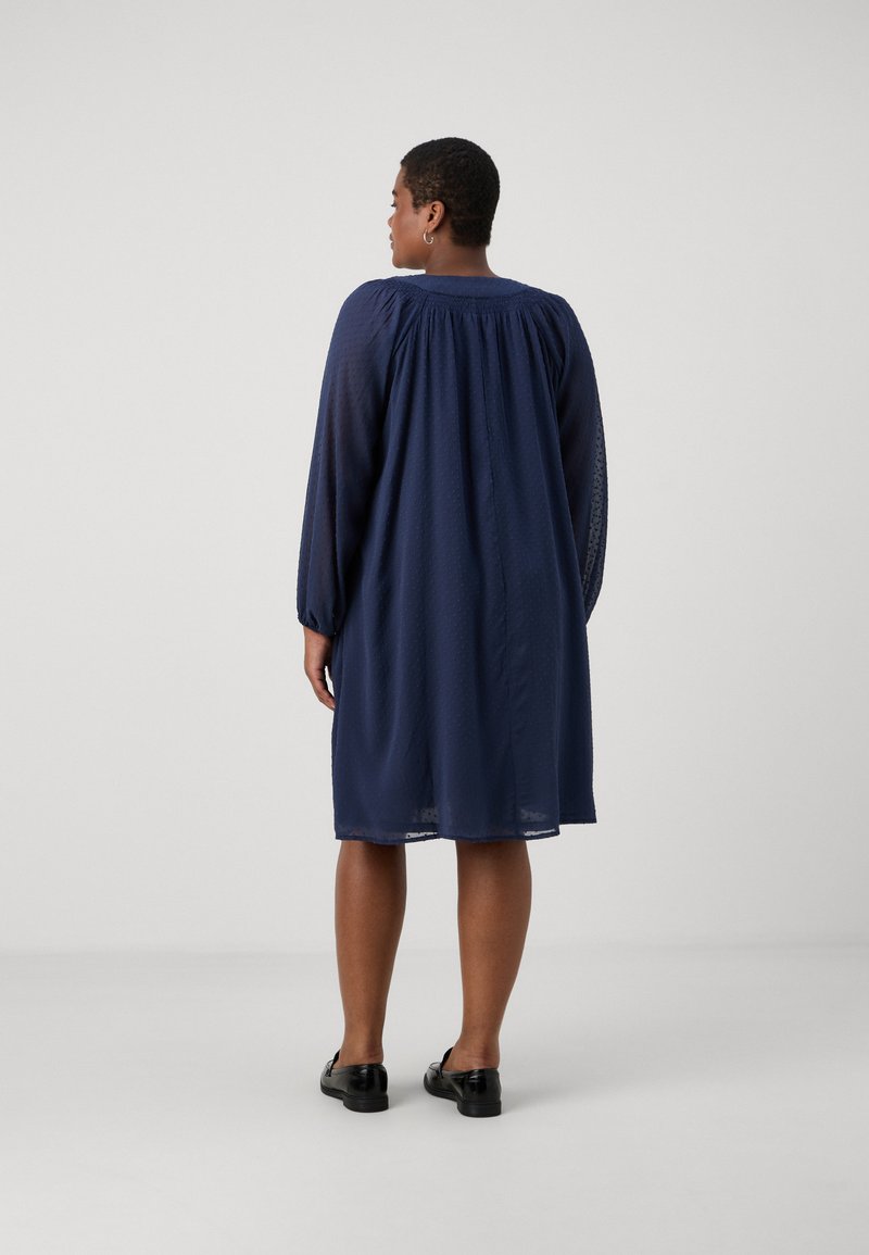 Robe bleu marine avec de longues manches transparentes et un col froncé. Tissu doux et texturé tombant juste au-dessus de la cheville. Chaussures noires à enfiler.