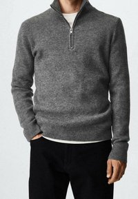 Grauer gerippter Strickpullover mit einem halbseitigen Reißverschlusskragen, langen Ärmeln und lockerer Passform, über einem weißen Hemd getragen, kombiniert mit schwarzen Hosen.