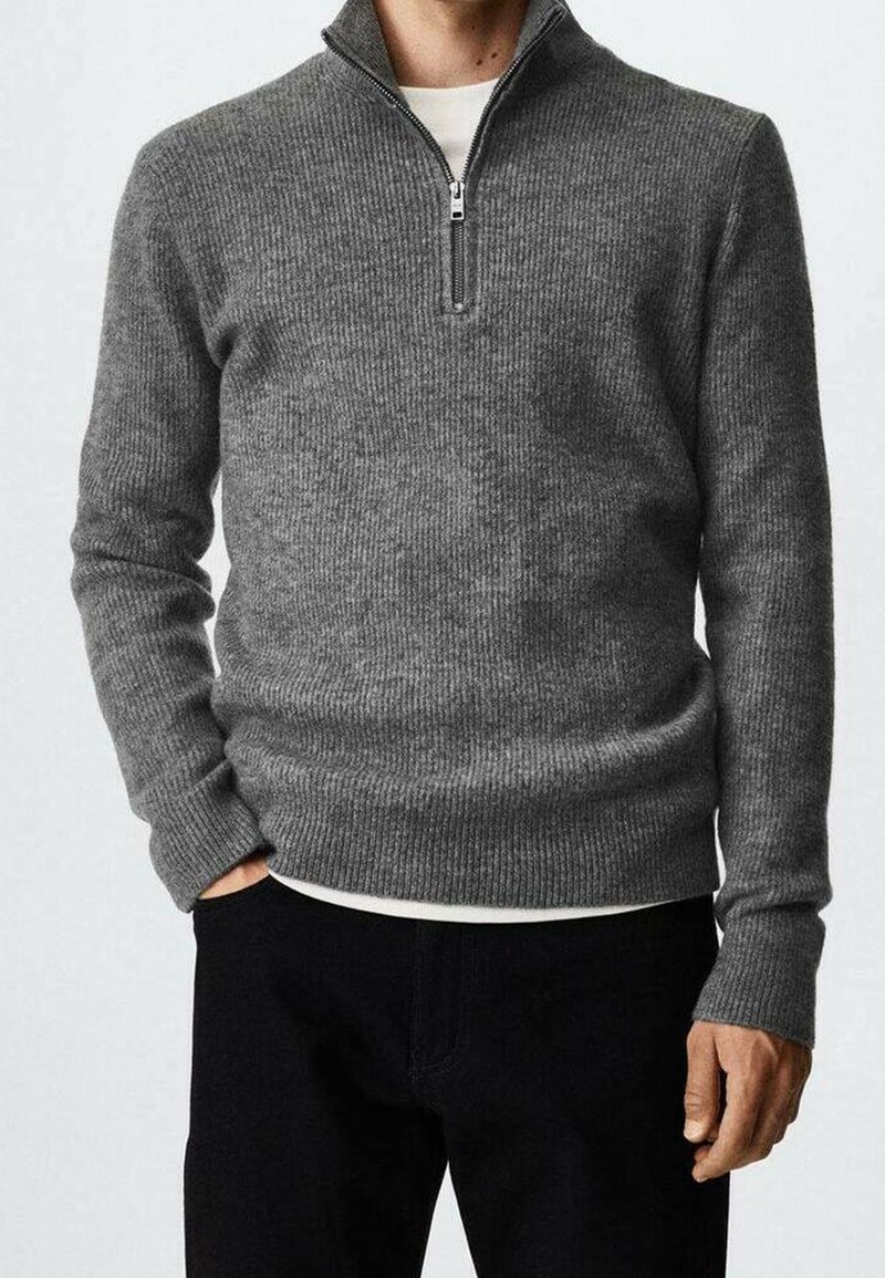Grauer gerippter Strickpullover mit einem halbseitigen Reißverschlusskragen, langen Ärmeln und lockerer Passform, über einem weißen Hemd getragen, kombiniert mit schwarzen Hosen.
