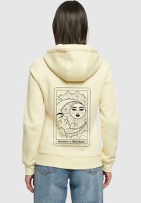 Gul huvtröja med en mjuk textur som har en svart tarotkortdesign p�å baksidan, som avbildar sol- och mån-bilder samt texten "Solälskare & Stjärnjägare."