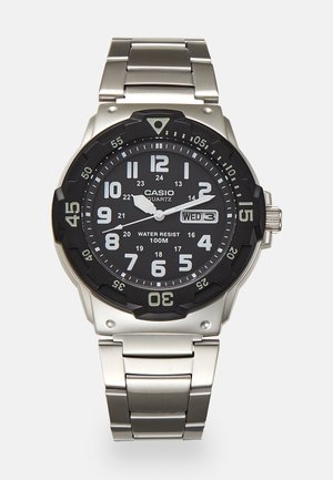 Relógio de quartzo metálico com mostrador preto, números brancos e display de data. Apresenta uma pulseira de aço inoxidável e resistência à água de 100m.