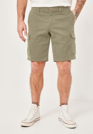 Man wearing olive green cargo shorts with side pockets, white high-top sneakers, and a white shirt, standing against a plain background.