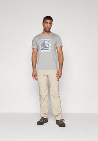 Camiseta gris con un gráfico azul central y texto, combinada con pantalones cargo beige. La vestimenta se complementa con botas de senderismo negras con cordones naranjas.