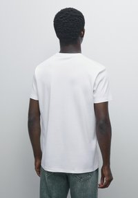Mand med mørk hud og krøllet hår står med ryggen til iført en ensfarvet hvid t-shirt med korte ærmer og blå jeans foran en grå baggrund.