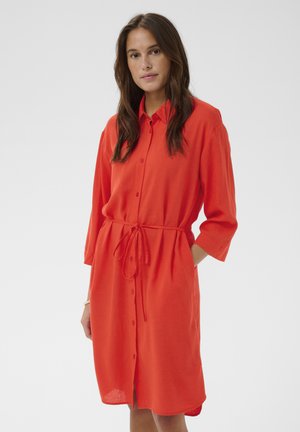 Kobieta z długimi brązowymi włosami, ubrana w jasnopomarańczową sukienkę na guziki z przewiązanym w talii paskiem i rękawami o długości do połowy przedramienia, stojąca na jednolitym tle.