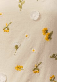 Tela crema con bordado floral colorido; presenta hilos amarillos, blancos y verdes en varias formas de flores y detalles intrincados.