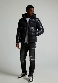 Black puffer jacket with a zip closure and hood, featuring a logo on the chest, paired with distressed black jeans and black sneakers.