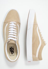 Sneaker in suede beige con suole in gomma bianca, design classico con lacci e strisce laterali bianche a contrasto. L'interno presenta un logo nero.