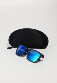 Sonnenbrille mit einem mattschwarzen Rahmen und blauen, verspiegelten Gläsern, neben einem schwarzen Hartschalenetui mit dezentem Logo platziert.