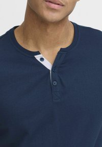 Námořnický modrý henley triko z měkkého materiálu, s bílou légou a dvěma modrými knoflíky a kulatým výstřihem.