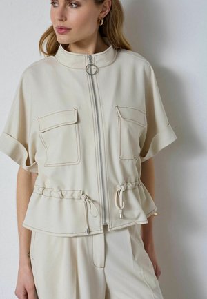 Mujer con chaqueta crema de manga corta con cremallera frontal, dos bolsillos en el pecho y cintura ajustable con cordón, combinada con pantalones a juego.
