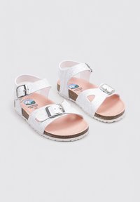 Sandalias blancas con correas texturizadas y acentos de hebilla metálica, con una plantilla rosa y una suela marrón y blanca.