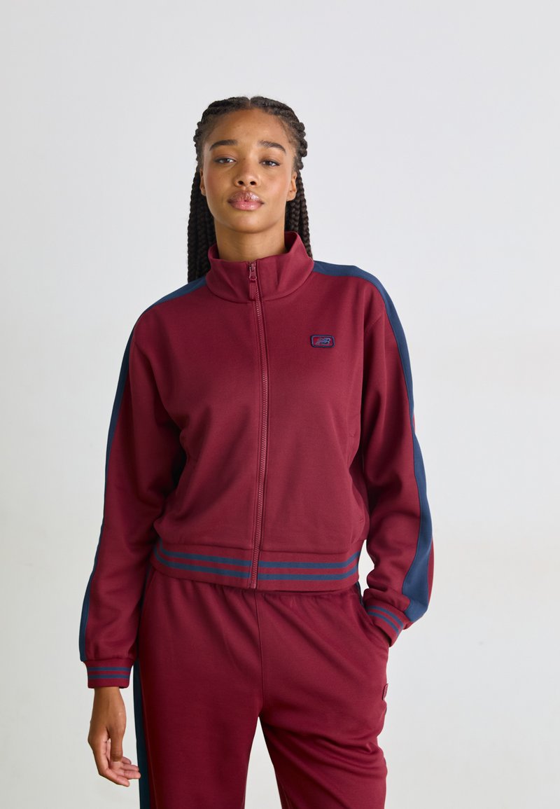 Bordeaux zip-jakke med høj krave, navy sidelameller og logo på brystet. Blødt stof, ribbede manchetter og talje. Matchende bukser.