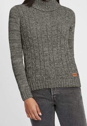 Femme portant un pull à col roulé en maille torsadée gris et un jean gris foncé, avec une petite pièce marron sur l’ourlet du pull.