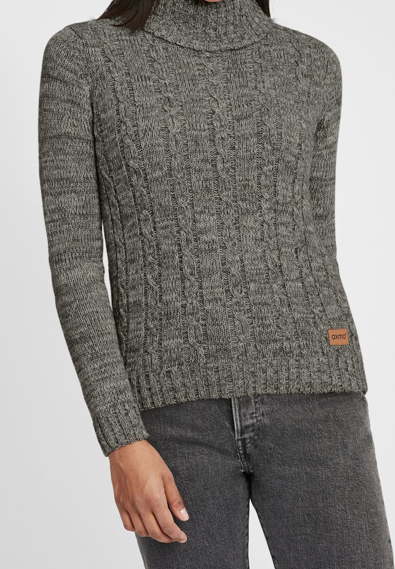 Femme portant un pull à col roulé en maille torsadée gris et un jean gris foncé, avec une petite pièce marron sur l’ourlet du pull.