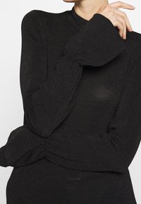 Haut noir à manches longues en tissu texturé avec un léger effet scintillant. Il présente un col montant et des poignets évasés, avec un détail froncé à la taille.