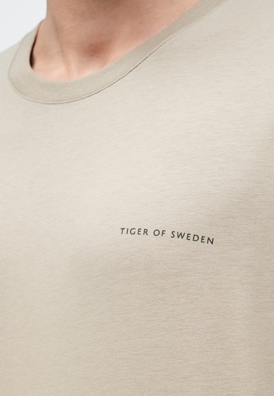 Bežs kokvilnas T-kreklis ar apaļu izgriezumu un ribotām malām. Priekšpusē kreisajā pusē ir melns uzraksts "TIGER OF SWEDEN". Gluda tekstūra.