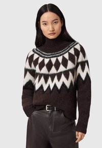 Maglione lavorato a maglia marrone con un motivo geometrico bianco e nero, colletto alto a coste e maniche lunghe, abbinato a pantaloni di pelle marroni.