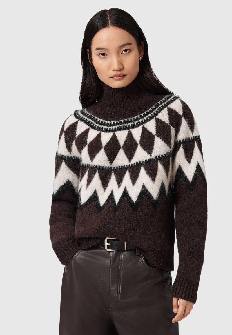 Maglione lavorato a maglia marrone con un motivo geometrico bianco e nero, colletto alto a coste e maniche lunghe, abbinato a pantaloni di pelle marroni.